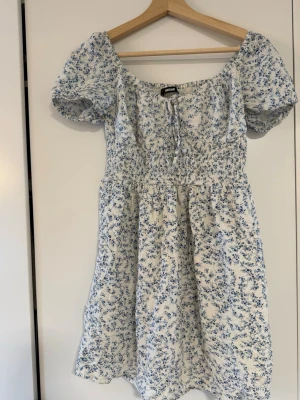 Blommig kort klänning från Bik Bok - Söt kort klänning från Bik Bok i vitt med blått blommönster. Klänningen har puffärmar, smock i midjan och knytning vid halsen. Perfekt för varma dagar och ger en romantisk vibe. Tillverkad i mjuk bomull som känns skön mot huden.