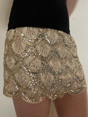 Kort paljettkjol i guld  - Supersnygg kort kjol täckt med guldiga paljetter i ett vågigt mönster på beige botten. Kjolen är perfekt för alla fester och tillställningar.