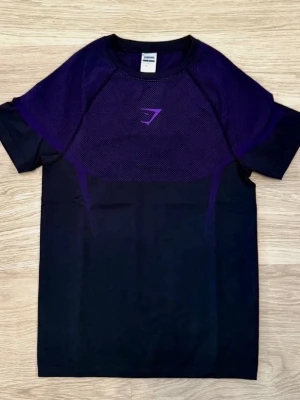 Svart och lila Gymshark t-shirt - Snygg svart t-shirt från Gymshark med lila meshdetaljer över axlar och bröst samt Gymshark-logga i lila framtill. Perfekt för träning med sitt lätta och ventilerande material. Kortärmad och modern passform.