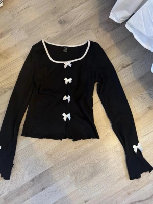 Svart cardigan med vita rosetter - Supersöt svart cardigan med långa ärmar och vita rosetter längs knäppningen och vid ärmsluten. Tröjan har vit kant runt halsen och en lätt croppad passform. Perfekt för en trendig och söt look.