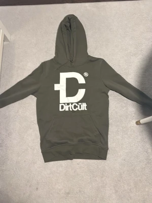 Olivgrön hoodie från DirtCult - Säljer en olivgrön hoodie från DirtCult med stor vit logga och text på bröstet. Hoodien har klassisk huva och magficka framtill. Tillverkad i mjukt bomullsmaterial som är skönt att bära. Perfekt för dig som gillar streetwear och vill ha en stilren look.