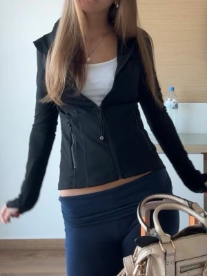 Svart träningsjacka med dragkedja - Snygg svart träningsjacka med figurnära passform och hög krage. Jackan har hel dragkedja framtill och två fickor med dragkedja. Perfekt för löpning eller gymmet. Materialet är stretchigt och följsamt. Lappen är bortklippt men tror den är storlek 2 eller 4