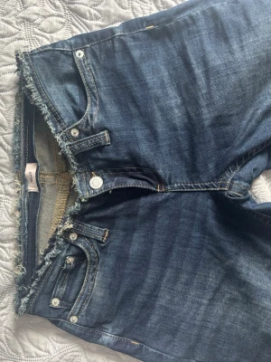 Mörkblå bootcut jeans med rå kant - Säljer ett par mörkblå bootcut jeans med rå, fransig midjekant och klassiska fem fickor. Jeansen har en låg midja och subtila slitningar för en avslappnad look. Perfekta för dig som gillar en edgy och trendig stil.