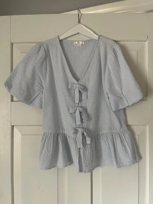 Blårandig blus med rosetter Gina Tricot - Söt blårandig blus från Gina Tricot med tre dekorativa rosetter framtill. Blusen har v-ringning, korta puffärmar och volang nedtill. Tillverkad i ett lätt, strukturerat tyg som känns luftigt och somrigt. Storlek 156/164 men passar även Xs. 