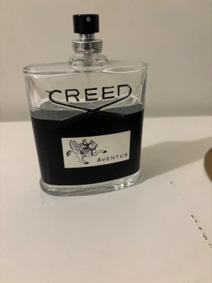 Creed Aventus parfym 100ml - Creed Aventus är en exklusiv parfym i en elegant, genomskinlig glasflaska med svart och silverdetaljer. Flaskan har en svart etikett med en silverhäst och ryttare samt texten 'Aventus'. Perfekt för dig som gillar lyxiga dofter. Locket är skadad men det viktigaste är den goda doften