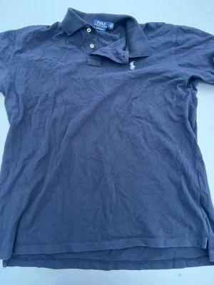 Ralph Lauren piké - SITTER SOM M,    Snygg marinblå pikétröja från Polo Ralph Lauren med klassisk krage, två knappar och vit broderad logga på bröstet. Tillverkad i mjuk bomull och har korta ärmar. Enkel att matcha till olika stilar.