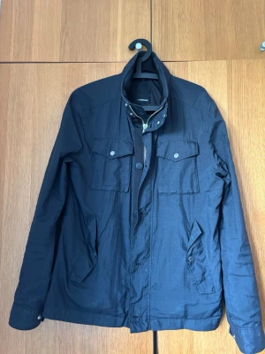  vindjacka från J.Lindeberg - Stilren vindjacka från J.Lindeberg med hög krage och tryckknappar. Jackan har två bröstfickor med lock och knappar samt två snedställda fickor framtill. Materialet är lätt och passar perfekt för blåsiga dagar. Diskret logga på ärmen.