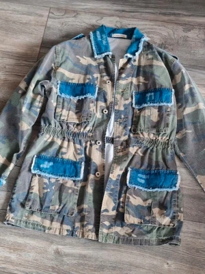 Camouflage parkajacka med blå detaljer - Som Ny !! Unik parkajacka i camouflage med fyra stora fickor och slitna blå denimdetaljer på fickor och krage. Jackan har lång ärm, knappar framtill och elastisk midja för skön passform. Perfekt för dig som vill sticka ut med streetstyle.