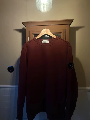 Vinröd tröja Stone Island - Vinröd stickad tröja från Stone Island med klassisk rund hals och lång ärm. Tröjan har en ikonisk svart patch med logga på vänster ärm. Enkel och stilren design, perfekt för dig som gillar streetwear och snygga detaljer. Hör gärna av dig, om du har några frågor. Ny pris 2999kr ✨