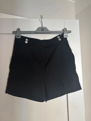 Svarta shorts med knappar och dragkedja - Stilrena svarta shorts med hög midja och två dekorativa guldknappar framtill. Klassisk passform med dragkedja och fickor i sidorna. Perfekta till sommaren för en clean och enkel look.