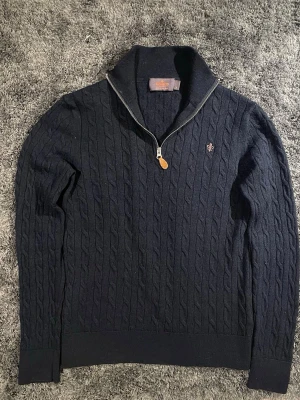 Mörkblå kabelstickad halfzip - Kabelstickad mörkblå tröja från Morris i 100% merinoull med halv dragkedja och hög krage. Tröjan har ett diskret broderat emblem på bröstet och ribbade muddar vid ärmslut och nederkant. Perfekt för dig som gillar klassisk och stilren design. Obs: tröjan har krympt i tvätten från S till en liten XS. Passar 155-165 max, tröjan sitter tajt runt en. Har du några frågor så är de välkomna! 