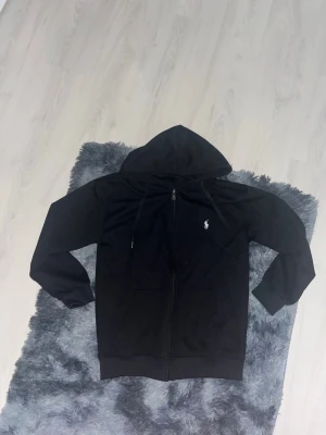 Svart hoodie från Polo Ralph Lauren - Svart hoodie från Polo Ralph Lauren med dragkedja och klassisk huva. Ikonisk vit logga broderad på bröstet och praktiska fickor framtill. Materialet är mjukt och bekvämt, perfekt för en avslappnad och stilren look. Finns 2st
