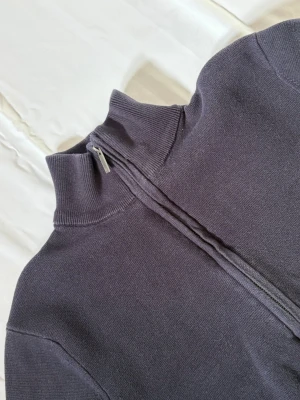 Mörkblå ziptröja från Bläck - Mörkblå stickad ziptröja från Bläck med hög krage och hel dragkedja framtill. Tröjan har ribbade muddar vid krage och ärmslut, vilket ger en clean och stilren look. Perfekt för lager på lager och enkel att matcha med olika outfits.