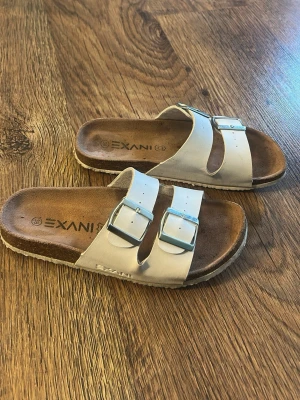 Vita sandaler med dubbla spännen Exani - Snygga vita sandaler från Exani med två breda remmar och silvriga spännen. Fotbädden är i brunt konstläder och sulan är platt med en lätt räfflad undersida. Perfekta för en avslappnad och trendig look.