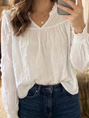 Vit blus med volang från Gina Tricot - Snygg vit blus från Gina Tricot med volangdetaljer längs ärmarna och smock vid ärmsluten. Blusen har v-ringning och är tillverkad i ett lätt, luftigt tyg som ger en avslappnad men ändå chic look. Perfekt att styla med jeans eller kjol.