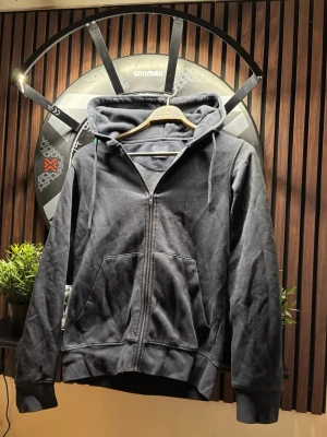 Svart Zip Hoodie - Klassisk svart hoodie med dragkedja från Jack & Jones. Hoodien har huva med dragsko, två stora fickor framtill och ribbade muddar vid ärmar och nederkant. Perfekt för en avslappnad och stilren look. Tillverkad i mjukt bomullsmaterial.