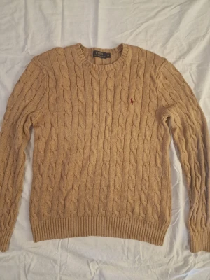 Polo Ralph Lauren kabeldtickad tröja - Kabelstickad beige tröja från Polo Ralph Lauren med rund halsringning och klassisk röd logga broderad på bröstet. Tröjan har långa ärmar och ribbade muddar vid ärmslut och nederkant. Perfekt för en stilren och avslappnad look.