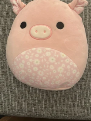 Rosa Squishmallows gris plysch - Supersöt rosa Squishmallows gosedjur i form av en gris med blommigt mönster på magen, små öron och knorr på svansen. Tillverkad i mjuk plysch och perfekt att krama eller ha som dekoration i rummet.