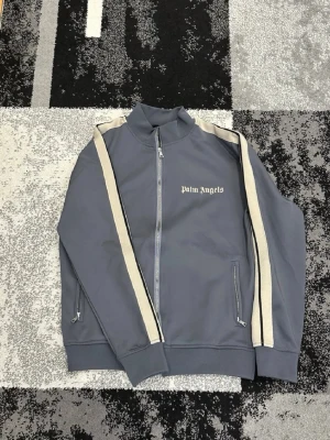 Grå ziptröja från Palm Angels - Snygg grå ziptröja från Palm Angels med vita kontrastband längs ärmarna och logga på bröstet. Tröjan har hög krage, dragkedja framtill och två fickor med dragkedja. Perfekt för en streetwear-look.