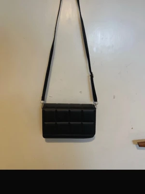 Svart quiltad axelväska - Snygg svart axelväska i rektangulär form med quiltat mönster. Väskan har bred axelrem och stängs med lock. Materialet ser ut att vara konstläder, vilket ger en modern och trendig vibe. Perfekt för dig som gillar stilrena accessoarer.