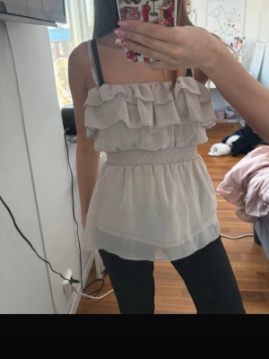 Ljusbeige volang topp med smock - Säljer en ljusbeige ärmlös topp med volanger över bysten och smock i midjan. Toppen har tunna axelband och ett luftigt, flowy tyg som ger en romantisk vibe. Perfekt för varma dagar eller när du vill känna dig extra söt.