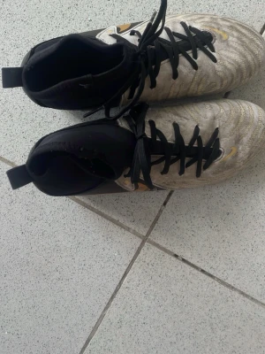 Nike fotbollsskor svart/guld - Säljer ett par Nike fotbollsskor med högt skaft i svart och guld. Skorna har snörning, svart ovandel och guldfärgade detaljer inklusive den klassiska Nike-loggan. Tillverkade i syntetmaterial för bra grepp och kontroll på planen.