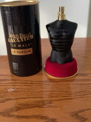 Jean Paul Gaultier Le Male parfym - Säljer en Jean Paul Gaultier Le Male parfym i den ikoniska flaskan formad som en manlig torso. Flaskan är mörkblå med svarta ränder och har ett silverfärgat sprayhuvud. Parfymen kommer med en rund röd sammetsbas med guldkant. Helt oandvänd fast utan prislapp. 