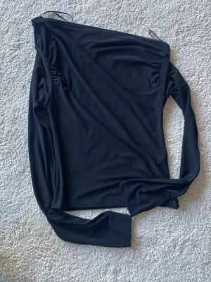 Svart oneshoulder topp Gina Tricot - Säljer en svart oneshoulder topp från Gina Tricot i mjukt stretchigt material. Toppen har lång ärm på ena sidan och är axelbandslös på andra, vilket ger en snygg asymmetrisk look. Perfekt att styla med jeans eller kjol för en trendig outfit. Fynda mer och spara på frakten 