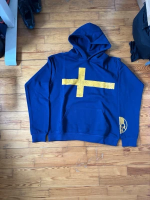 Blå hoodie med gul korsdesign - Snygg blå hoodie med stor gul korsdesign på framsidan och tryck på ärmen. Baksidan har texten 'Peace In War' och detaljerad text om Sveriges historia. Hoodien har huva och magficka, perfekt för en unik och stilren look.