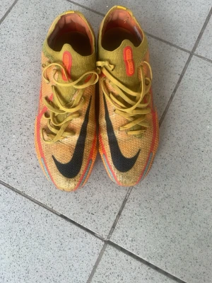Nike Phantom GT2 gula fotbollsskor Elite - Säljer ett par Nike Phantom GT2 fotbollsskor i gult med svarta och röda detaljer. Skorna har en låg profil, snörning och stora svarta Nike-loggor på ovansidan och sidan. Ovandelen är i syntetmaterial med textur för bättre bollkontroll. Perfekta för dig som vill sticka ut på planen.