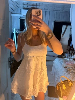 Vit kort sommarklänning med volang - Söt vit kort klänning med tunna axelband som knyts på axlarna. Klänningen har volangdetaljer och en lätt rynkad struktur i tyget. Perfekt för varma dagar och har en luftig, flowy passform.