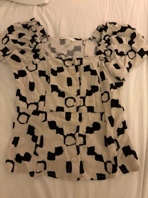 Mönstrad kortärmad blus med knappar - Snygg blus i beige med svart abstrakt mönster och puffiga korta ärmar. Blusen har knappar framtill och en lätt figursydd passform. 