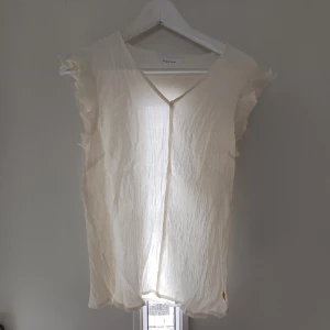 Kräm vit blus med volangärmar från Coster - Luftig kärm vit blus från Coster i tunn bomull med v-ringning och volangdetaljer vid ärmarna. Storleken stämmer inte på bilden eftersom den krympte i tvätten så rekommenderar att handtvätta.