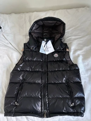 Svart dunväst från Moncler med huva - Snygg svart dunväst från Moncler med glansig finish och vadderad design. Västen har en stor huva, två dragkedjefickor framtill och klassisk Moncler-logga på bröstet. Perfekt för lager-på-lager och streetstyle. Det finns även en fraktpåse från moncler men den är hos min mamma