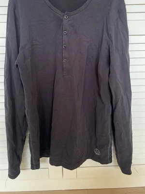 Mörkgrå långärmad topp East West - Mörkgrå långärmad topp från East West med rund halsringning och knappdetaljer framtill. Tillverkad i mjuk bomull som känns skön mot huden. Enkel och stilren design som passar till det mesta.