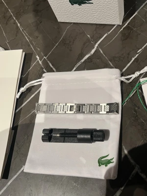 Silverfärgad klocklänk från Lacoste - Säljer en stilren silverfärgad klocklänk från Lacoste i rostfritt stål. Länken har ett diskret mönster och klassisk design med viklås. Perfekt för dig som vill byta ut eller uppgradera din klocka. Verktyg för att justera längden medföljer.