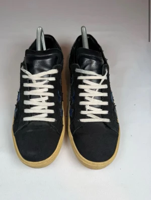 Saint Laurent skor svarta! - Snygga svarta sneakers från saint lauren  med blå glittriga stjärnor på sidorna och vita skosnören. Skorna har en beige sula och är tillverkade i mocka och läder. Perfekta för dig som vill sticka ut med coola detaljer och unik design. Stl 41