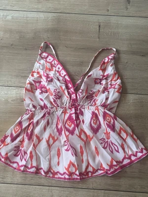 Mönstrad linnetopp med smala axelband - Snygg topp med färgglatt rosa och orange mönster på vit botten. Djup v-ringning, smala axelband och resår i midjan. Aldrig använd. Passar även s.