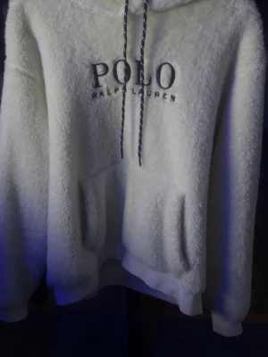 Vit teddy  Ralph Lauren hoodie - Supermjuk och fluffig beige hoodie från Polo Ralph Lauren med stor broderad logga på bröstet. Tröjan har huva med snörning och en stor magficka. Perfekt för kyliga dagar när du vill vara både bekväm och snygg.