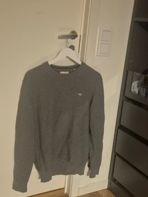 Grå tröja från GANT - En stilren grå tröja från GANT med rund halsringning och diskret logga på bröstet. Tröjan har lång ärm och ribbade muddar vid ärmslut och nederkant. Perfekt för lager-på-lager och enkel att matcha med jeans eller byxor.