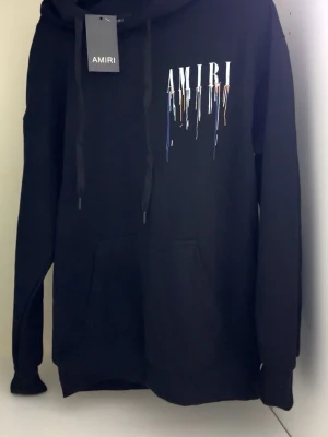 Svart hoodie från Amiri med tryck fraction - Svart hoodie från Am,iri med stor vit logga och färgstänk i blått, orange och vitt både fram och bak. Klassisk känguruficka, huva med dragsko och ribbade muddar. Snygg streetwear vibe och riktigt skön passform. Perfekt för dig som gillar exklusiva märken och unik design.