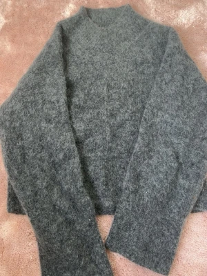 Grå fluffig mohairtröja från H&M - Mysig grå stickad tröja från H&M i mohair- och ullblandning. Tröjan har en hög hals och långa ärmar, perfekt för kalla dagar. Den fluffiga strukturen ger en trendig och avslappnad look.