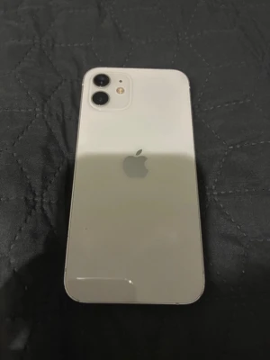 Vit iPhone 12 med dubbla kameror - Säljer en vit iPhone 12 med stilren design och rundade hörn. Baksidan är i glas och har två kameror samt Apple-loggan. Framsidan har en stor skärm med tunn ram och Face ID. Perfekt för dig som vill ha en snygg och modern smartphone.