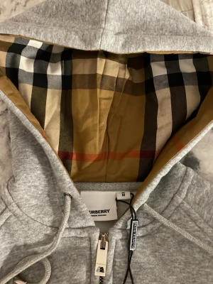 Grå zip-hoodie från Burberry - Snygg grå hoodie från Burberry med dragkedja och klassiskt rutigt foder i huvan. Hoodien har fickor framtill, ribbade muddar och justerbar dragsko i huvan. Perfekt för dig som gillar stilrena och exklusiva plagg med en twist.