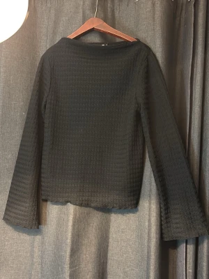 Svart långärmad topp med struktur - En svart långärmad topp med diskret strukturerat tyg och rak passform. Toppen har en enkel, stilren design och är perfekt att matcha med både jeans och kjol. Materialet känns lätt och något stretchigt.