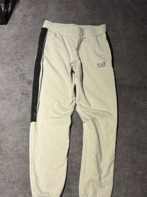 Gråa EA7 mjukisbyxor med svarta detaljer - Snygga gråa mjukisbyxor från EA7 Emporio Armani med svarta detaljer längs sidorna och logga på vänster ben. Byxorna har resår i midjan och muddar vid bensluten. Perfekta för en avslappnad och trendig look.