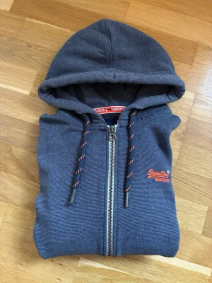 Superdry hoodie blå grå zip Nyskick - Blågrå hoodie från Superdry med dragkedja, huva och dragsnören med röda detaljer. Broderad Superdry-logga i orange på bröstet och mjukt foder på insidan. Klassisk passform och ribbade muddar vid ärmar och nederkant. Perfekt för en avslappnad streetwear-look. Knappt använd så den är som ny.