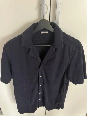 Mörkblå frottéskjorta med kort ärm - Avslappnad mörkblå skjorta i mjuk frotté med korta ärmar och klassisk krage. Skjortan har knappar framtill och en avslappnad passform, perfekt för en chill och trendig look. Strl L. 