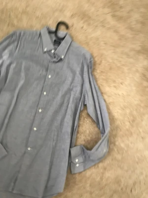 Ljusblå skjorta med knappar - Klassisk ljusblå skjorta med lång ärm och vita knappar. Skjortan har en smal krage och är tillverkad i ett tunt, mjukt bomullstyg som passar perfekt till jeans eller chinos. Enkel och stilren design utan mönster eller extra detaljer.