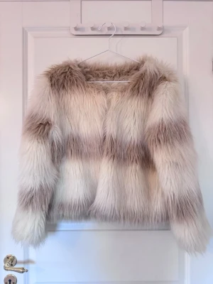Fluffig beige och vit fuskpälsjacka - Säljer en superfluffig fuskpälsjacka i beige och vit med breda ränder. Jackan har rak passform och rundad halsringning, perfekt för dig som vill sticka ut med en trendig vinterlook. Stängs framtill utan synliga knappar eller dragkedja. Den har tyvärr ett litet hål under armen på sidan men den syns knappt till med pälsen över🩷🩷